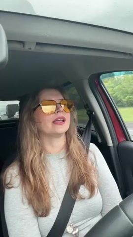 A woman is driving a car. She is wearing a gray long-sleeved shirt, a seatbelt, sunglasses with orange lenses, and a necklace with a heart pendant. She is smiling and talking as she drives, occasionally gesturing with her right hand. Other vehicles are visible in the background, suggesting she's on a highway. The weather appears to be overcast or rainy. The interior of the car is visible, including the rearview mirror and the passenger seat area.