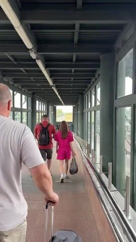 A person is walking down a covered walkway with a suitcase. A couple is walking ahead of them. The walkway has large windows on one side, offering a view of train tracks.
