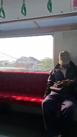 A man wearing a cap, jacket, and pants sits on a red seat on a train and looks at his phone. The train car has white walls and ceiling, a window, and green handles hanging from the ceiling. Outside the window, there are houses and a telephone pole.
