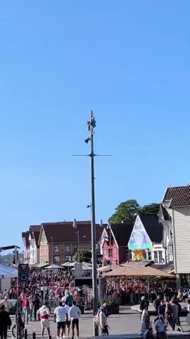 A bright, sunny day with a clear blue sky. The scene appears to be a waterfront area, possibly a harbor or a town square adjacent to the water. In the foreground, a tall pole with multiple devices attached to it, including cameras and what appear to be speakers or lights, stands prominently. Several people are walking around in the foreground, some casually strolling, others engaged in conversation. One person is seen riding a bicycle. In the mid-ground, a large crowd of people is gathered, many wearing pink attire. There are also tents and stalls set up, suggesting a market or an event. Colorful banners and decorations are visible, further indicating a festive atmosphere. In the background, several traditional-looking buildings with pitched roofs line the waterfront. Beyond the buildings, a large cruise ship is visible on the water, along with several sailboats. The overall impression is one of a lively and populated public space on a pleasant day.