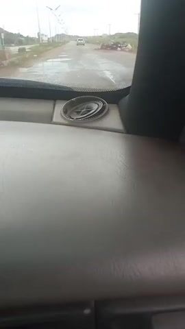 The video shows a view from inside a vehicle, looking out through the windshield. The dashboard is visible in the foreground, a grey, textured surface with a circular speaker grille mounted on it. Beyond the windshield, a paved road stretches into the distance, lined with streetlights on both sides. A white car is visible driving away from the camera in the center of the road. The environment appears to be a somewhat developed area, possibly a highway or a main road, with some construction or debris visible on the right side of the road. The sky is overcast. The camera is stationary, indicating the vehicle is in motion.