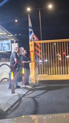 Two men stand by a yellow metal gate at night. One man, wearing a dark shirt and gray pants, holds a phone or device. The other man, wearing a dark shirt and jeans, stands to his left. An Israeli flag is visible on a flagpole behind them. A security camera is mounted on a building to the left. The gate appears to be closed.