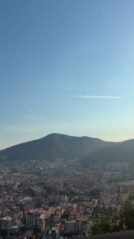 The video shows a panoramic view of a coastal city nestled at the foot of a mountain. The sky is clear and blue with a faint streak of clouds. The city is densely populated with buildings featuring red-tiled roofs, interspersed with taller, modern structures. A bay or inlet is visible on the left, with the calm sea stretching to the horizon. The mountain, covered in greenery, dominates the background. The perspective suggests the viewpoint is from a higher elevation, overlooking the cityscape. The overall impression is one of a sunny day in a Mediterranean or similar climate.