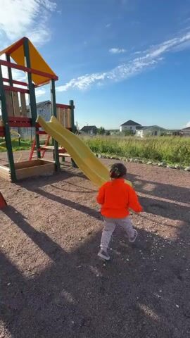 A child wearing an orange jacket and gray pants walks towards a yellow playground slide on a sunny day. The child then climbs up the slide and slides down. The playground is made of wood and plastic, with a sandpit and a climbing wall. The ground is covered with gravel. In the background, there are houses and trees. The sky is blue with some white clouds.
