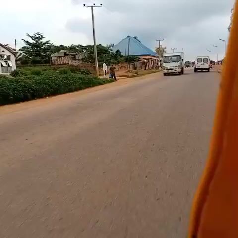 The video shows a road with a white truck driving down it. On the side of the road, there is a person standing next to a blue bucket. There are also trees and buildings in the background. The sky is cloudy. The camera appears to be mounted on a moving vehicle.