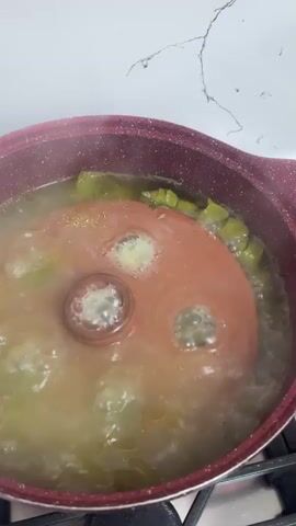 A close-up shot shows a pot of food being cooked on a stovetop. The pot is filled with a light-colored liquid and appears to contain pieces of green vegetables. A red lid, with a central knob and three visible steam vents, is placed on top of the pot. Bubbles rise to the surface of the liquid and escape through the vents in the lid, indicating that the food is boiling. The pot is a dark red color and has two handles. The stovetop is visible at the bottom of the frame, with a black burner knob. A white countertop with a marbled pattern is visible in the background. The overall scene depicts a cooking process in a kitchen environment.