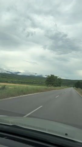The video captures a scenic drive through a rural landscape in Biaga, Bulgaria, from the perspective of a moving vehicle. An asphalt road with white lane markings stretches into the distance, flanked by green fields and lush vegetation, including a prominent tree on the left side of the road. In the background, verdant mountains are partially shrouded by low, wispy clouds, suggesting an overcast day. The sky is filled with grey clouds. The overall scene depicts a natural, hilly environment under a cloudy sky. No people or visible text are present in the video.