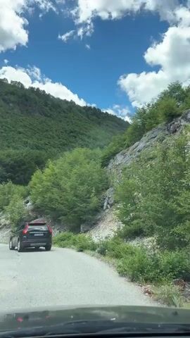 A dark-colored SUV drives on a paved road curving to the left, ascending a steep, heavily wooded mountainside. The road is bordered by dense green foliage and rocky outcrops. The sky is a vibrant blue with scattered white, puffy clouds. The car is the only vehicle visible. The camera appears to be inside another vehicle, looking forward and slightly to the right. The time stamp is 00:00 to 00:04.