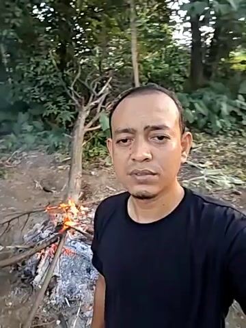 A man is standing in front of a campfire in a wooded area. The man is wearing a dark-colored T-shirt. The fire is small, with flames and embers visible. The ground around the fire is littered with ash and bits of wood. In the background, there are trees and greenery. The man looks directly at the camera. The camera appears to be hand-held.