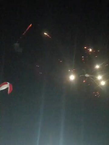 The video captures a nighttime scene with a dark sky illuminated by fireworks. In the upper left quadrant, a bright burst of fireworks is visible, displaying orange and purple sparks. To the right of this, a bright, circular light, possibly the moon or a strong light source, shines. A few smaller, scattered lights, possibly more fireworks or distant lights, are also present in the sky. In the lower left corner, a portion of a red and white object, possibly a flag or banner, is visible. The lower part of the frame shows vertical light streaks, indicative of camera movement or light sources from the ground. The overall atmosphere suggests a celebration or event taking place outdoors at night.
