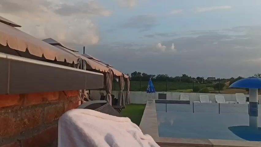 This media file captures a serene outdoor scene at what appears to be a resort or recreational area, likely in Sopot Urban Municipality, Serbia. The time of day is dusk, with the sun setting in the background, casting a warm glow across the sky and illuminating the clouds. The weather seems pleasant, with a mix of sunshine and dramatic cloud formations.

The primary subject of the scene is an outdoor swimming pool, its surface reflecting the sky and surrounding elements. Deck chairs are neatly arranged around the pool, some covered with white sheets, suggesting the end of the day or preparation for closure. A distinctive blue mushroom-shaped umbrella stands in the pool. Beyond the pool area, there's a grassy lawn dotted with more chairs and umbrellas, a covered seating area, and some trees. In the distance, houses and vegetation are visible, indicating a rural or semi-rural setting. The overall mood is peaceful and tranquil, evoking a sense of relaxation as the day concludes.

There are no people visible in the frame. The objects present include the swimming pool, lounge chairs, umbrellas (one blue mushroom-shaped, others potentially stored), covered seating areas, a towel draped over a brick structure in the foreground, and some distant structures that might be buildings or playground equipment. The lighting suggests either early morning or late evening. The presence of lounge chairs and a swimming pool points to a leisure facility. No visible text is present in the media file.