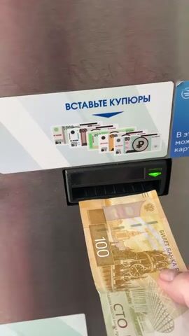 The video shows a person inserting a 100 ruble banknote into a ticket machine. The machine has a slot where banknotes can be inserted, and a sign above the slot that says "Insert banknotes" in Russian, with a diagram of different banknotes that the machine accepts.  The machine briefly displays a red light, and then ejects the bill. The person then reinserts the bill.  The machine is silver colored and appears to be made of metal. The person's hand is visible in the video.