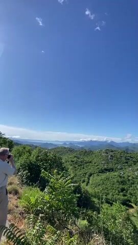 A person is seen from the chest down, holding a smartphone with both hands, appearing to capture a photo or video of a vast natural landscape. The phone screen shows the scene being recorded. The viewpoint is elevated, overlooking rolling green, forested mountains and valleys. In the far distance, a large body of water (likely a lake or sea) is visible, with more mountain ranges beyond. The sky is bright blue with a few scattered white clouds, indicating clear, sunny weather during the daytime. The overall impression is a scenic panoramic view from a high vantage point in a mountainous region. The location is Gornja Brova, Montenegro.