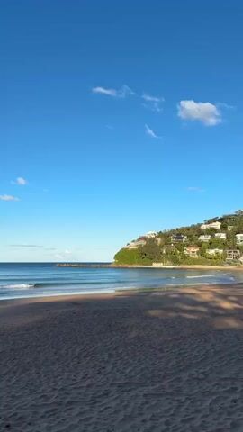 The video pans across a sandy beach with gentle waves rolling onto the shore under a clear blue sky. Lush green hills dotted with houses rise behind the beach, and in the distance, a headland is visible. The lighting suggests it is either early morning or late afternoon, casting long shadows across the sand. There are no people or animals visible in the frame.