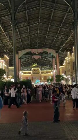 A group of people are walking through a covered area with a high, industrial-looking ceiling supported by large pillars. The area is illuminated by warm, yellow lights on the buildings lining the sides. The buildings appear to be designed in a historical style, with ornate architecture and decorated with strings of lights. A younger child in a striped shirt and pants is walking away from the camera in the foreground, while other people are walking towards and away from the camera in the midground. Some individuals are carrying bags or holding onto children. The overall atmosphere seems to be that of a bustling public space, possibly an entertainment venue or a themed area.