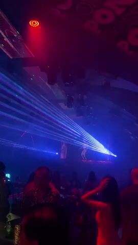 This media file depicts a vibrant and energetic indoor event, likely a club or concert venue, characterized by dynamic lighting and a palpable sense of celebration. The scene is dominated by an array of stage lights, including powerful blue spotlights and intense laser beams that crisscross the space, creating a mesmerizing visual spectacle. These lights illuminate a raised platform or stage area where a large screen displays abstract visuals or perhaps a performer's silhouette.

The subjects in the foreground are silhouettes of people, suggesting a lively crowd gathered to enjoy the event. They appear to be standing or seated, engaged with the performance unfolding before them. The atmosphere is dark, with the primary illumination coming from the stage lighting, casting a cool blue and purple hue over the surroundings. The overall mood is one of excitement and festivity.

While the specific time of day is not discernible, the indoor setting and the nature of the event imply it is likely an evening or nighttime affair. There are no clear indications of weather conditions. The location context provided is Khan Daun Penh, Cambodia, suggesting this event is taking place in that region.

Notable details include the intricate patterns of the laser lights and the fog or smoke that catches the light, adding depth and atmosphere. The stage design incorporates a tiled or brick-like texture at the bottom, contrasting with the smooth, illuminated screen above. The video captures a sense of movement and immersion in the event, drawing the viewer into the energetic ambiance. No visible text is present in the media file.