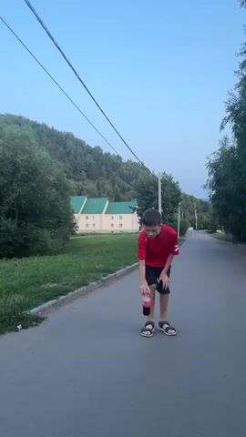 A young boy in a red shirt and black shorts is standing on a paved road, outdoors. He is holding a yo-yo in his right hand and appears to be in the process of dropping it. The yo-yo is suspended in the air, just above the road surface. Behind him, the road stretches into the distance, with trees and hills lining either side. In the background, a building with a light-colored facade and green roofs can be seen amongst the trees. The sky is clear and blue.