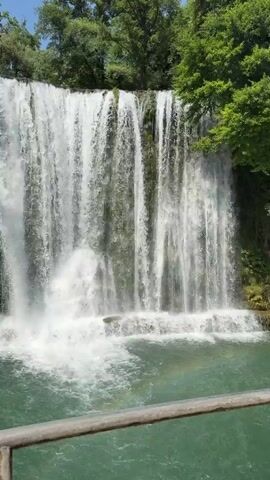 The media file showcases a beautiful, wide waterfall in Jajce, Bosnia and Herzegovina. The waterfall cascades down a moss-covered rock face into a clear, turquoise pool below. Lush green trees frame the top of the waterfall, suggesting a natural and serene setting. The sun is shining, creating dappled light on the water and highlighting the spray from the falls.

A metal railing is visible in the foreground, indicating that there is likely a viewing platform or walkway. In the final seconds of the clip, a person wearing a blue cap is partially visible at the bottom left, suggesting visitors are present to enjoy the view. The overall mood is peaceful and awe-inspiring, capturing the raw beauty of nature. The scene is likely taking place during the daytime, under fair weather conditions, given the bright sunlight and clear skies.