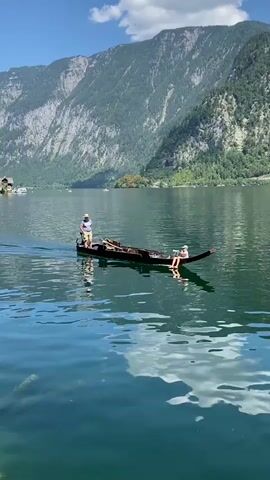 A long, narrow wooden boat is being paddled across a large body of water. The boat is dark brown and has a pointed bow and stern. There are two people in the boat. One person, wearing a white hat and a light-colored shirt, stands at the stern and appears to be paddling with a long oar. Another person, wearing a light-colored hat and a light-colored shirt, sits near the bow, facing forward. The boat is moving from left to right in the frame. The water is a deep blue-green with reflections of the sky and clouds. In the background, a large, steep mountain covered in green trees rises from the water. The sky is blue with scattered white clouds. The overall scene is serene and picturesque.