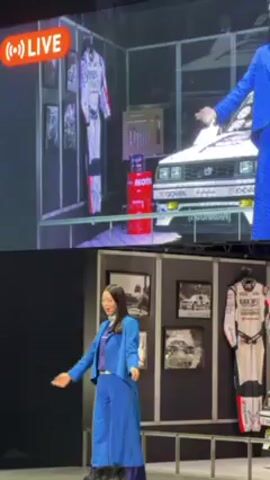 A presenter in a blue pantsuit stands on a stage. Behind her, on display, are photos of cars and a racing jumpsuit. A large screen shows a live feed of a car show with many attendees. The presenter gestures with her right hand toward the screen. She points her left hand in the air. The stage is simple, with a black backdrop and a metal railing.