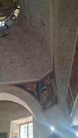 The video shows the interior of a church. The camera pans upwards, revealing a dome with frescoes. A large section of the dome's fresco has deteriorated and fallen away, exposing the stone structure beneath. A large, ornate chandelier hangs from the ceiling. Below the dome, an iconostasis, a wall of icons and religious imagery, is visible. Light streams in from windows, illuminating the interior. There are no people visible in the frame. The architecture suggests an Orthodox Christian church.