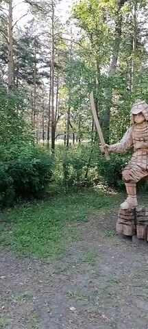 The video shows a wooden sculpture of a samurai warrior in a forest. The samurai is standing on a base made of logs, holding a sword in its right hand. The sculpture is detailed, showing the samurai's armor and helmet. The background is a forest with trees and foliage. The video appears to be filmed from a slight distance, showing the entire sculpture and its surroundings.