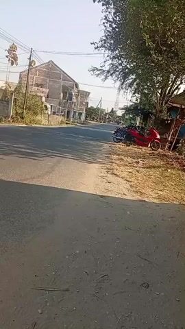 The video shows a paved road with dry grass and dirt on the side. Two motorcycles are parked on the side of the road. A building with multiple stories and exposed beams is visible in the background. There are trees and power lines also present in the scene. The lighting suggests it is daytime.