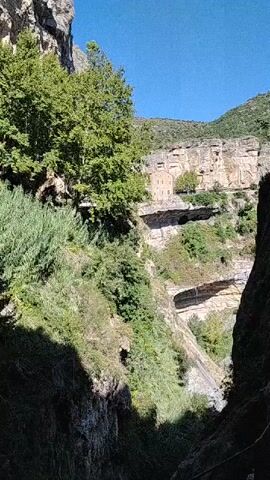 This video captures a scenic view of a gorge with lush vegetation and a building nestled into the cliff face. The camera pans downwards, revealing a waterfall cascading into a pool of water. The environment is natural and somewhat wild, with rocky cliffs and dense greenery. The building appears to be ancient and integrated with the natural landscape, possibly a monastery or hermitage. The sunlight illuminates the scene, highlighting the textures of the rocks and foliage.