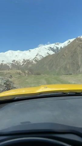 The video shows a view through the windshield of a yellow vehicle, with a black dashboard and steering wheel visible. The foreground shows the vehicle interior. In the background, a wide grassy field stretches toward large, snow-capped mountains under a clear blue sky. The vehicle appears to be moving forward on a dirt path. At one point, feathers hang down momentarily from the rearview mirror.