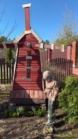 The video shows a young child standing next to a red wooden windmill in a garden. The child is wearing a beige jacket, green pants, grey boots, and a beige hat. They are smiling and waving at the camera. The windmill is in front of a wooden and brick fence, and there are various trees and plants in the background. The sky is blue and clear. The child is positioned in front of a patch of earth in which several small plants appear to be growing. The scene is well lit and appears to be taking place during daytime.