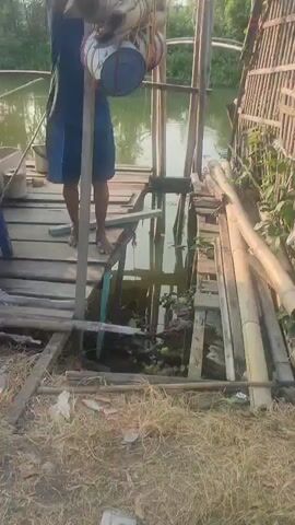 A person is standing on a wooden platform, operating a device that appears to be a water pump. They are wearing a blue t-shirt and blue shorts. The platform is next to a body of water, possibly a river or canal, with vegetation on the banks. There are several bamboo poles supporting the structure. The ground in front of the platform is covered in dry grass and some debris.