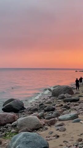 The video shows a beach scene at sunset. The sky is a gradient of pink and orange. The sea is calm and reflects the colors of the sky. The beach is covered in sand and large rocks. Two women are walking along the shoreline. They are both wearing dark clothing. One woman is facing away from the camera and looking at the water. The other woman is facing the camera and smiling. There is some vegetation and a small structure visible in the background.