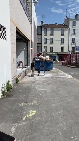 The video captures a daytime scene in what appears to be an alleyway or service entrance behind a building. A man, wearing a dark t-shirt and light-colored pants, is sweeping the asphalt ground with a broom. He is standing near a large, blue industrial dumpster filled with trash, including cardboard boxes and various debris. A small blue rolling cart is positioned in front of him on the ground.

To the man's left is the side of a building with a large open loading bay door, a white wall, and a visible ventilation grate. A red fire extinguisher is mounted on this wall. In the background, across the alleyway, are multi-story buildings with light-colored facades, numerous windows, and balconies, characteristic of urban European architecture. One building prominently displays a sign that reads "HARRY'S COFFEE," and another sign partially visible reads "SIECLE." The sky is clear and blue, suggesting sunny weather. The ground shows some white and yellow dust or markings. The man continues to sweep the area throughout the video.