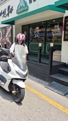 A person wearing a helmet, vest, and gloves sits on a white scooter parked in front of a building with green and white signage. The person is looking down and holding a phone. The building appears to be a restaurant. The scooter has a large black storage box on the back. The environment is outdoors with a sunny sky.