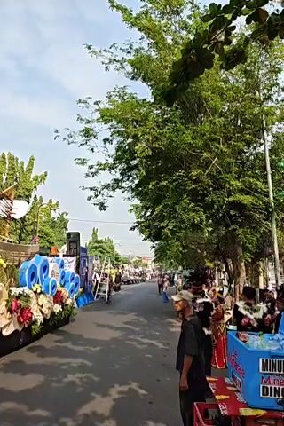 The video shows a street parade with several decorated floats. The first float is adorned with large blue and white circular objects, flowers, and fabric. The second float is decorated with various fruits and vegetables, including what appear to be eggplants, bananas, and gourds, with people dressed in traditional attire. The third float features a large, white, curved structure made of what looks like plastic cups or bowls. People are standing on the side of the street, watching the parade. There are trees and buildings visible in the background. The weather appears to be sunny.