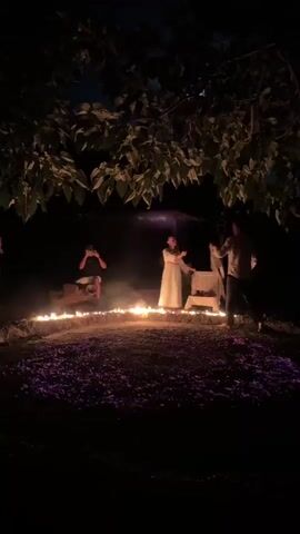 A group of people are gathered around a bonfire at night, lit by numerous small candles. The ground is covered with purple petals. Two people in white robes are dancing. One person is playing an accordion and another is sitting. There are trees and foliage above the scene. The atmosphere is dark and mystical.