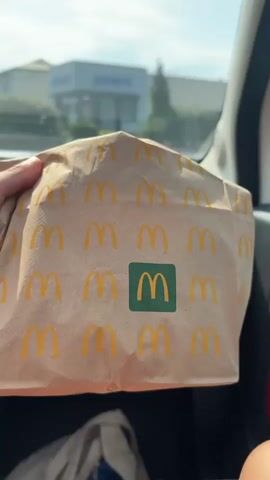The video begins with a close-up shot of a brown paper McDonald's bag held by a person's hand. The bag has the McDonald's logo printed repeatedly on it, with a larger logo in the center. The background is blurred, showing a building and a bright sky. The perspective then shifts to a view from inside a car, looking out the window. We see the side mirror reflecting a person, likely the driver, with blonde hair and sunglasses. Outside the car, a drive-thru lane is visible with green painted areas and white dashed lines on the asphalt. Several buildings are in the background, including what appears to be a McDonald's restaurant with a yellow facade and red signage. There are trees and a street light. The sky is clear and blue with some power lines visible. The car is moving, as indicated by the changing perspective and the slight motion blur. The video ends with the car continuing through the drive-thru lane.