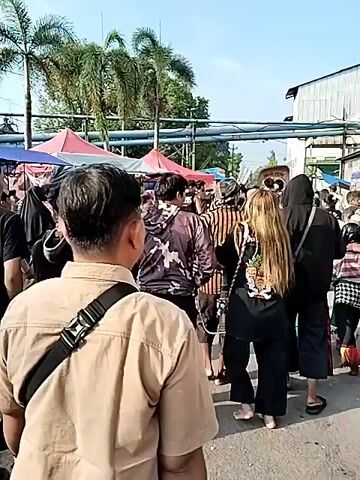 The video depicts an outdoor gathering or event, likely a procession, under a bright, sunny sky.

The initial frames (0:00-0:01) show a crowd of people, mostly adults, walking casually in an area with several pop-up tents with red and blue tarps. Palm trees and overhead power lines are visible in the background. People are dressed in casual attire, including t-shirts and shorts. Some partially visible text on a sign suggests "VIDAL."

The scene then transitions (0:01-0:05) to a more focused view of a procession. People walk along a paved surface past temporary market stalls or kiosks, characterized by blue tarps. A prominent figure is a woman in the foreground (0:03-0:05) wearing traditional Javanese attire: a brown and black horizontally striped top and a white and black batik-patterned skirt. She carries a large, intricately designed banner or sign. To her left, a man in a light-colored security-style uniform carries a red and white Indonesian flag. Other participants in the procession include men and women, some wearing traditional clothing or hijabs, and one person carrying a yellow flag. The overall atmosphere suggests a cultural or civic event. The large banner held by the woman features elaborate graphics but no clearly discernible text from the video angle.