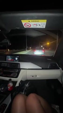 The video is a first-person point-of-view shot from inside a car at night. The dashboard is visible, with the infotainment screen displaying information. A person's legs, wearing shorts, are in the foreground, with their feet resting on the dashboard. The car is moving, as indicated by the motion blur of the road and the streetlights outside. Another car is visible in front, its taillights illuminated. The interior of the car appears to be a luxury model, possibly a BMW, given the dashboard design and the airbag warning sticker visible on the sun visor. The overall atmosphere is casual and relaxed, suggesting a nighttime drive.