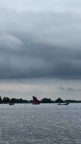 The video shows a wide shot of a body of water under a cloudy sky. Several boats are visible on the water, including a sailboat with a red sail, a smaller boat in the distance to the left, and another boat with a person in it to the right. There are also what appear to be two birds or ducks in the water near the boat on the right. The shoreline in the background is lined with trees. The water is rippling, suggesting a light breeze. The overall mood is calm and perhaps a bit overcast.
