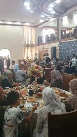 This is a video of a gathering or event inside a large indoor space, possibly a banquet hall or reception venue. The room is well-lit by overhead lighting. In the foreground, a table is laden with food, and several people, mostly women and children, are seated around it, eating and interacting. The people are dressed in various attire, with many of the women wearing headscarves. In the background, a group of musicians is performing on a raised stage area, playing instruments and singing. The overall atmosphere appears to be festive and celebratory, with a mix of dining and entertainment.