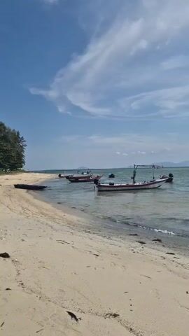 The video shows a beach with boats anchored in the water. The beach has light-colored sand, and there are a few trees on the left side of the frame. Several boats, of varying sizes and designs, are anchored in the shallow water near the shore. The water is clear and calm, with the sun reflecting off its surface, creating a shimmering effect. The sky is blue with scattered white clouds. In the distance, a low mountain range is visible. The camera slowly pans from left to right, providing a panoramic view of the beach and the sea.