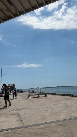 The video shows a view of a concrete pier on a sunny day with a bright blue sky and scattered clouds. A white, corrugated roof overhangs the left side of the frame. Several people are visible on the pier. On the left, a person wearing a green shirt and black shorts is standing and looking down. Further down the pier, a person in a blue hijab and white pants is walking. To the right, a metal railing with some equipment is positioned on the pier. The ocean is visible in the background with waves gently lapping against the shore. The overall atmosphere appears to be relaxed and casual.