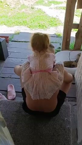 The video shows a person from behind, seated on a wooden porch. The person is wearing black shorts. A young child, possibly a toddler, is sitting on the person's shoulders, facing away from the camera. The child is wearing a light pink dress with white lace trim. The porch is covered in a gray rug, and a pair of pink slippers are visible to the left of the person. Green grass and some wooden structures are visible in the background. The lighting suggests it is daytime.
