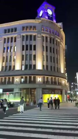 The video shows a street scene in Japan at night. The camera looks up at a large building with a clock tower. The clock tower is lit up with blue light and has the word "SEIKO" on it. The building is also lit up with white lights. People are walking across a crosswalk in front of the building.