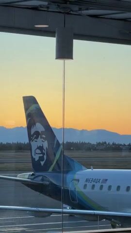 A close-up shot from inside an airport terminal shows the tail of an Alaska Airlines plane, emblazoned with the company's distinctive smiling face logo, against a backdrop of a sunset. The plane is parked on the tarmac, with its registration number N634QX visible on the fuselage. In the distance, another small plane is seen on the runway, and a line of trees and mountains forms the horizon. The warm hues of the sunset cast a soft glow over the scene, creating a peaceful and serene atmosphere. The shot is framed by a window, with a light fixture hanging from the ceiling adding a touch of industrial design to the natural beauty of the sunset.