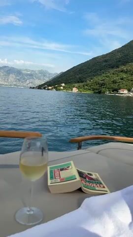 The video shows a close-up view of a glass of white wine and two books on the deck of a boat. The books appear to be paperbacks with red and green covers. In the background, there is a body of water, likely a bay or a lake, with mountains on the far side. The sky is blue with some clouds. The water is calm and reflects the sky and the surrounding landscape. The scene is set during the daytime, with bright sunlight. The camera is positioned low, looking out over the water. There are no people visible in the video. The overall impression is one of relaxation and leisure.