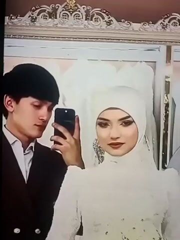 A young couple is taking a selfie in front of a mirror. The man is on the left, wearing a suit, and is holding the phone. The woman is on the right, wearing a white wedding dress with a hijab. Behind them, there are other wedding dresses visible in the reflection of the mirror. There is loud, festive music playing.