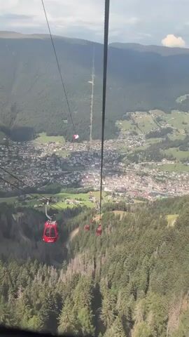 This is a first-person perspective video taken from inside a cable car as it ascends a mountain. Below, a dense forest covers the mountainside, transitioning into a village nestled in a valley. The village consists of numerous buildings with red roofs, interspersed with green fields and roads. In the distance, the valley opens up to more mountainous terrain under a cloudy sky. Several other red cable cars are visible on the cable line, ascending alongside the viewer's car. The viewer's cable car appears to be one of the closest to the camera. A cable runs vertically down the center of the frame, with a red and white marker attached to it. The camera is angled downwards, providing a clear view of the descending landscape.