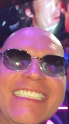 The video opens with a close-up of a smiling bald man wearing sunglasses, with a gold chain visible.  Behind him is a purple illuminated sign with the letters "BPEX." The camera shifts to show a DJ setup with two DJs. One DJ is wearing sunglasses and a black jacket. The second DJ is in a suit jacket looking at their phone. Large images of faces are displayed on screens behind them. The setting appears to be a nightclub.